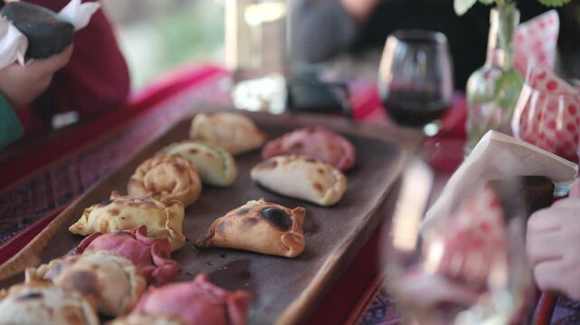Hand selecting a freshly baked Argentine empanada from a rustic serving platter filled with assorted pastries during a shared meal with friends and family, slow motion close-up shot.