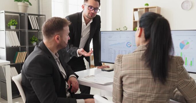Manager with analysts leads strategy meeting presentation on graphs. Three colleagues view graphs on a computer monitor and plan steps for growth in an office. Clear goals and strong teamwork.