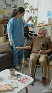 Vertical video of young female nurse checking IV drip of senior patient and talking to senior man on sofa. Ideal for nursing home and geriatric care marketing campaigns
