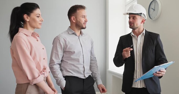 Couple with agent and inspector in hardhat. They review details during apartment viewing with a broker and contractor, considering purchase and investment. Clear real estate discussion concept.