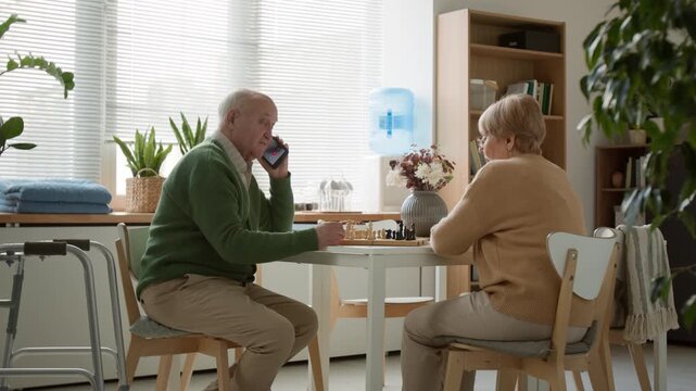 Side view shot of senior man talking on smartphone while playing chess with senior woman in retirement home. Video for senior care or retirement lifestyle marketing