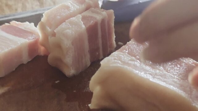 A high-quality macro close-up video capturing a person precisely slicing a piece of fresh raw pork belly into thick slabs using a sharp kitchen knife on a wooden cutting board