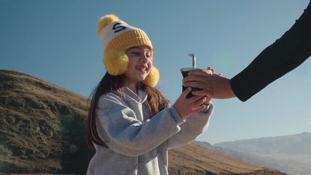 Young girl reaches for a traditional yerba mate gourd while enjoying a family moment in a mountainous Andean landscape, close-up outdoor shot, family bonding and leisure lifestyle concept