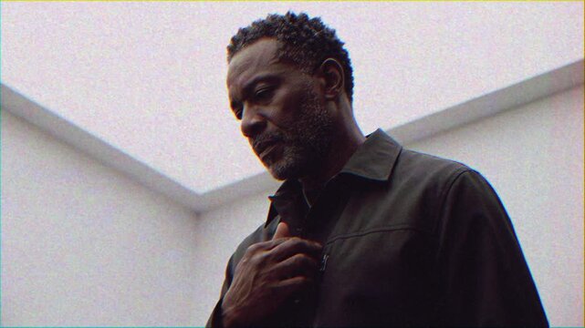 Black man under skylight adjusting necklace, veteran actor reflecting in minimalist studio with overhead skylight, soft grainy