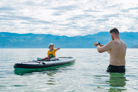 Father photographing toddler on paddleboard in mountain lake