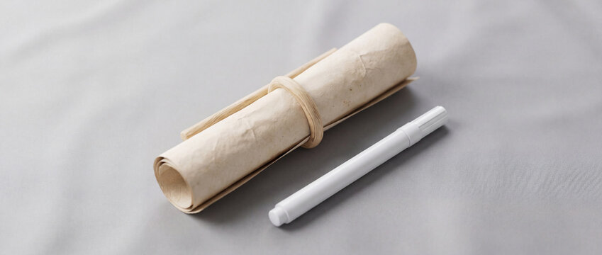 A rolled parchment paper scroll tied with a natural cord lying next to a white modern pen on a clean grey fabric background