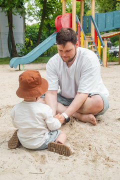 Father and toddler playing in a sandy playground with colorful slides and climbing structure in a green park, summer family leisure outdoors.