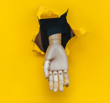 A wooden hand protrudes through a torn hole in yellow paper, begging for alms. Concepts of prosthetics, disability, and poverty.