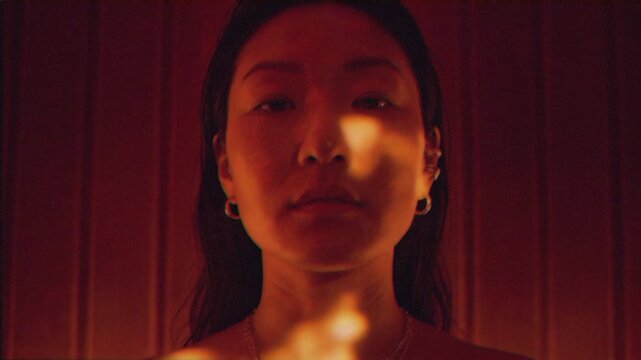 Asian woman closeup in warm light, wooden panel background with candlelike glow, closed eyes then slow opening, subtle sweat