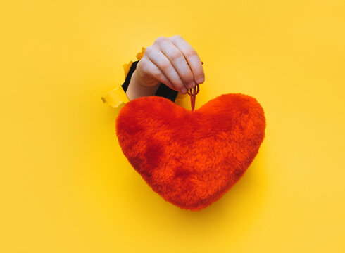 A child's hand holds a red plush heart through a torn hole in yellow paper. Concept of love and Valentine's Day.