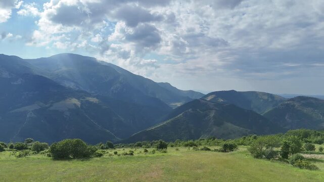 Paesaggio montano con creste sovrapposte illuminate da raggi di sole filtrati tra le nuvole, prato verde in primo piano e atmosfera naturale profonda e luminosa