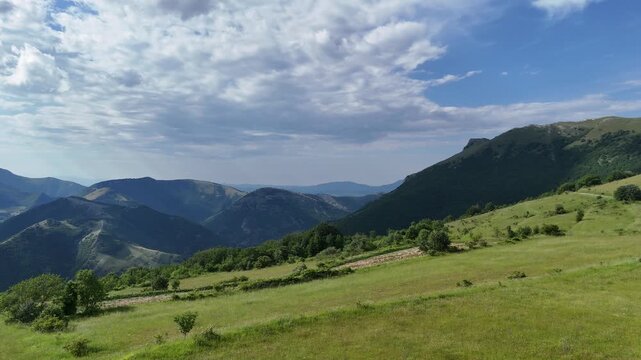 Ampio prato verde ai piedi di una montagna e cielo azzurro punteggiato da nuvole bianche. Scena naturale luminosa e tranquilla