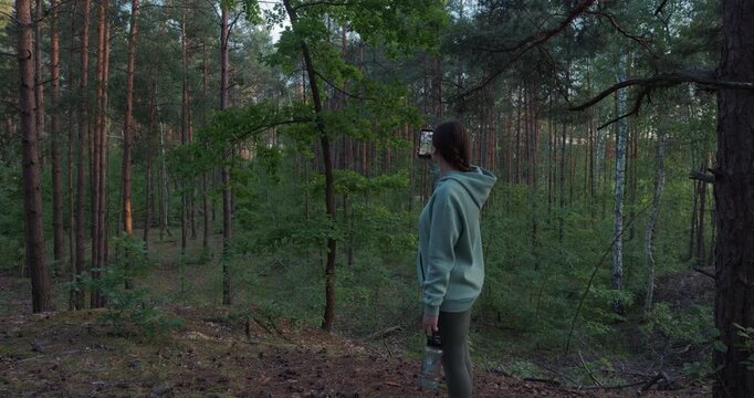 Young woman holding smartphone and taking photo in dense pine forest
