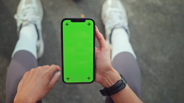 Top view of Asian woman holding smartphone with green screen and tracking markers during workout, female athlete pointing finger at mobile phone mockup on street for fitness app and technology concept