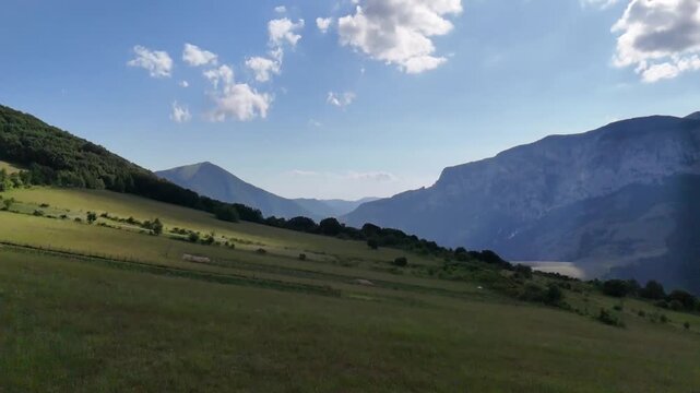 Ampio prato verde ai piedi di una montagna e cielo azzurro punteggiato da nuvole bianche. Scena naturale luminosa e tranquilla