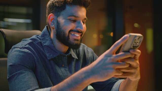 Happy Indian business man browsing mobile phone in evening office at table joyful Arabian businessman smiling scrolling web app Hindu male guy professional smile looking in smartphone at night agency