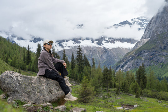 The female tourist sits on a rock, with snow-capped mountains and a forest in the background