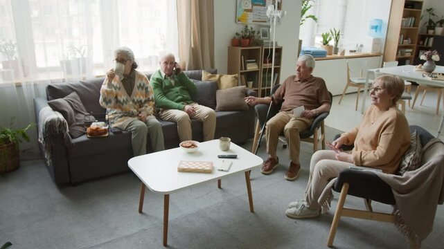 Wide angle shot of seniors watching TV together and relaxing in assisted living facility common room, drinking tea and socializing. Perfect for senior care marketing and retirement community promo