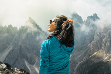 Close up profile woman hiker in turquoise fleece holding hair looking at dramatic mist covered mountain peaks background Cadini di Misurina Dolomites Italy summer. © Evaldas