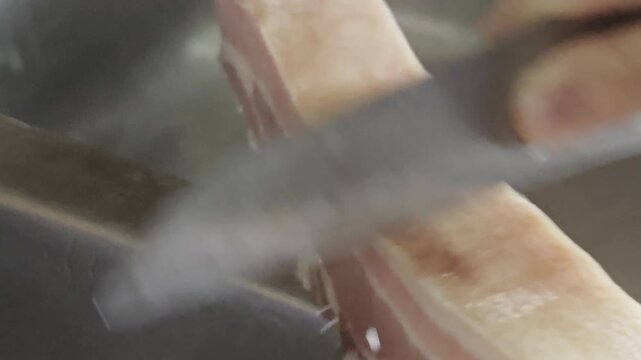 A high-quality close-up video capturing a person meticulously scraping the skin rind of a fresh, raw pork belly slab using a sharp chef's knife over a stainless steel kitchen sink.