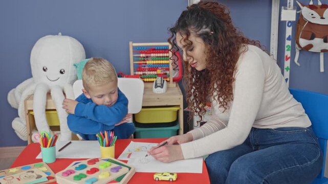 Woman teacher drawing with pencil at small table while little boy rests chin on arm and watches in preschool building; learning calm.