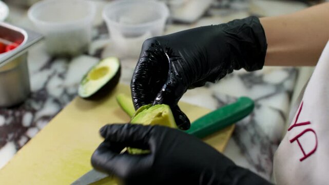 Hands in black gloves slicing and peeling avocado on a cutting board in kitchen prep