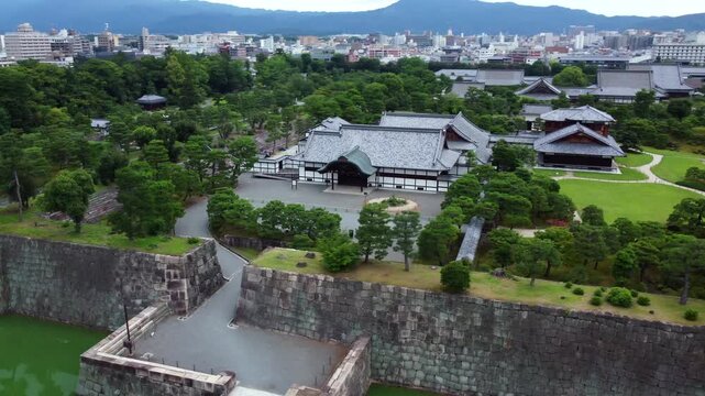Drone orbits above Nijo Castle complex revealing traditional Japanese rooftops, stone moat walls, and manicured grounds with city skyline beyond.