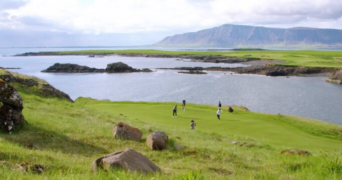 A group of golfers play on a lush green course located on a scenic coastline, with a vast mountain range across the water under a partly cloudy sky.