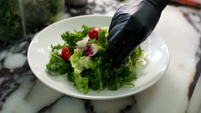 Gloved hand adds cherry tomatoes to fresh green salad on marble kitchen counter