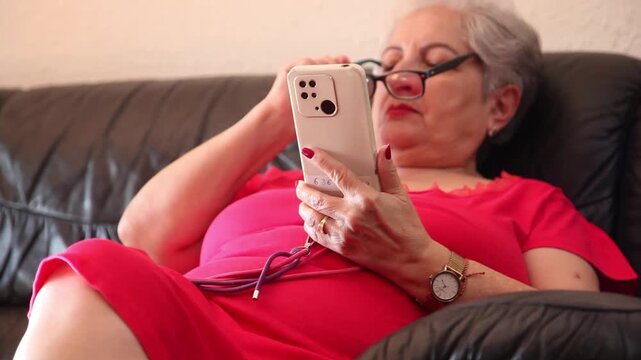  Mujer mayor con bata rosa usando un tel&eacute;fono inteligente en el sof&aacute; de su casa. Mujer mayor con gafas navegando por el tel&eacute;fono inteligente en casa. Senior activo, estilo de vida tecnol&oacute;gico moderno.