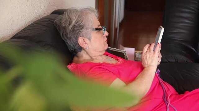  Mujer mayor con bata rosa usando un tel&eacute;fono inteligente en el sof&aacute; de su casa. Mujer mayor con gafas navegando por el tel&eacute;fono inteligente en casa. Senior activo, estilo de vida tecnol&oacute;gico moderno.