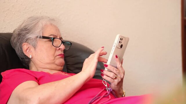  Mujer mayor con bata rosa usando un tel&eacute;fono inteligente en el sof&aacute; de su casa. Mujer mayor con gafas navegando por el tel&eacute;fono inteligente en casa. Senior activo, estilo de vida tecnol&oacute;gico moderno.