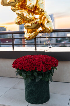 Large bouquet of red roses in box and gold heart shaped balloons on rooftop terrace at sunset