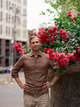 Smiling handsome man standing with hands on hips near blooming pink rose bush on city street