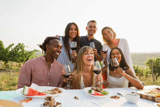 Happy adult friends having fun drinking red wine and eating together with vineyard in background - Multiracial people doing party at summer time - Main focus on center girl face - UGC content