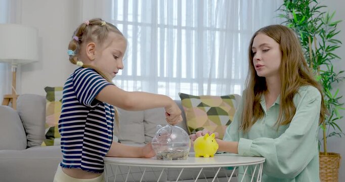 Caucasian single mother watches preschool daughter sort coin beside clear and yellow piggybank, coaching saving habit, money counting, responsibility, and warm parent child learning at home.