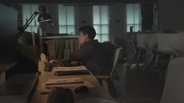 A businessman sitting at a classic wooden desk in a dark office at night leaning forward while working on a computer under warm lamp light.
