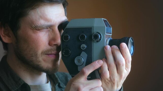 Filmmaker with a beard looking through the viewfinder of a vintage 8mm movie camera and then lowering it, smiling with satisfaction after shooting a scene indoors with natural light