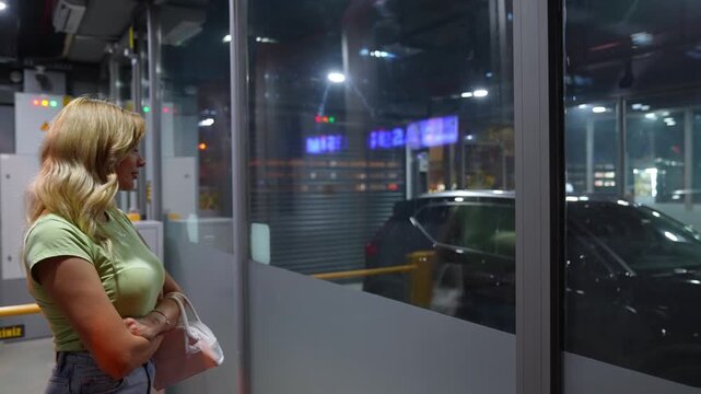 A young woman waits behind a glass partition inside a smart parking facility while her vehicle is being brought to the pickup area. The scene reflects automated parking service, user experience and mo