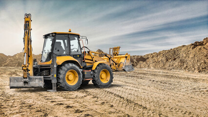 Fototapeta premium An excavator loader operates on a construction site moving soil and materials during daylight. The background shows piles of earth and a clear sky
