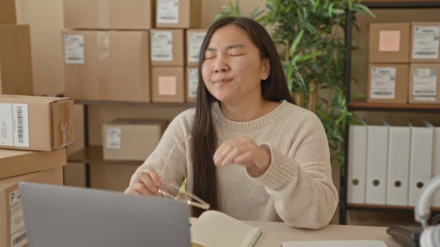 Woman at a packing desk with laptop and cardboard parcels pinches nose and removes glasses while sorting orders in building; work stress.