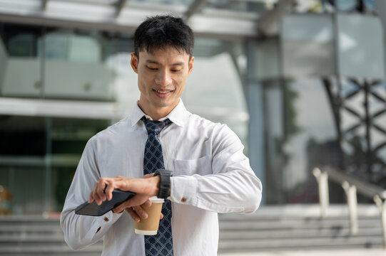 Asian businessman office worker checking time on watch while holding coffee cup outside the building