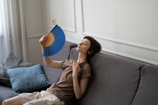 Exhausted woman sitting on sofa fanning herself struggling with sweltering weather, feel heat dehydration. Airless apartment with one window, health deterioration, vulnerability to hot weather, no AC.