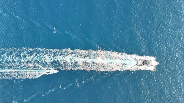 Aerial view of two boats gliding through the calm blue sea, leaving white wake trails behind them as they navigate the tranquil waters under bright sunlight. Summer vacation concept. Ocean shoreline
