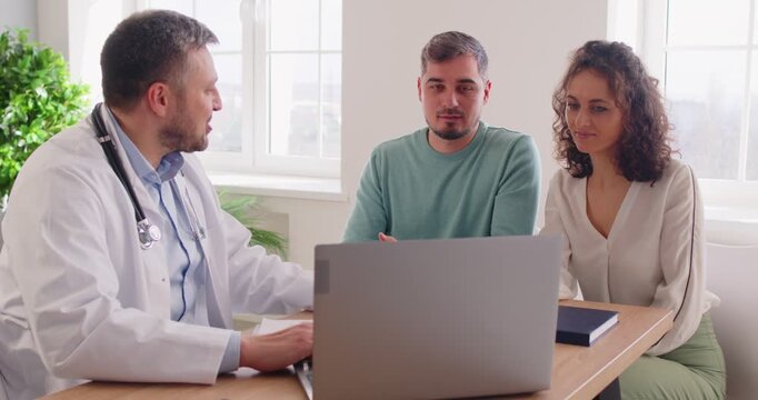 Friendly gynecologist warmly shakes hands with couple, sharing good news of their pregnancy. Happy husband and wife talking to doctor while sitting in front of laptop in modern clinic office.