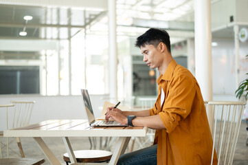 Fototapeta premium Asian man freelancer working on a laptop while sitting at table in comfortable cafe environment.