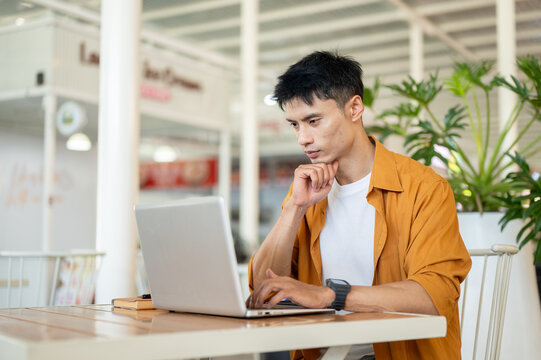 Asian man focused on browsing online with laptop while sitting at table in a contemporary cafe.
