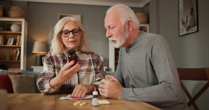 Two older adults sit together at a table in a home. One person examines a bottle of medication while the other listens. There are small packets and another bottle in front of them.