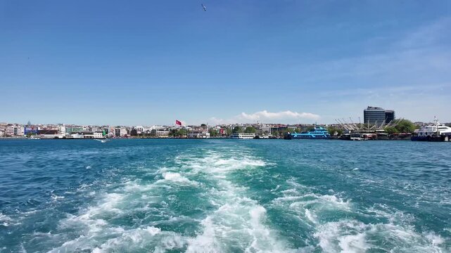 White foam and waves trace behind a moving ferry on the Bosphorus. Seagulls flying over the sea with city coastline and Turkish flag in the background.