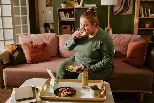 Young plus size Caucasian woman eating sandwich on sofa while using tablet at home, illustrating sedentary snacking and screen time. Ideal for wellness, nutrition, obesity awareness marketing